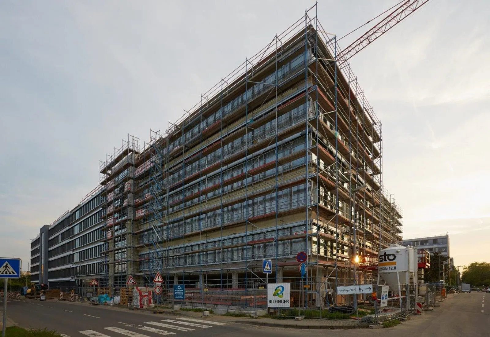 Verwaltungsneubau in der Dämmerung, vollständig eingerüstet, Kran über der Fassade und abgesperrte Baustellenzufahrt im Vordergrund